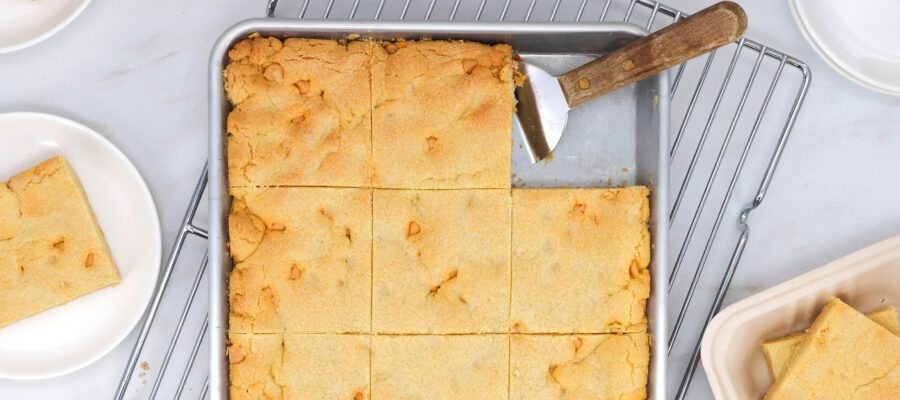 Golden brown cornbread squares arranged in a baking pan with a wooden-handled spatula nearby on a cooling rack.