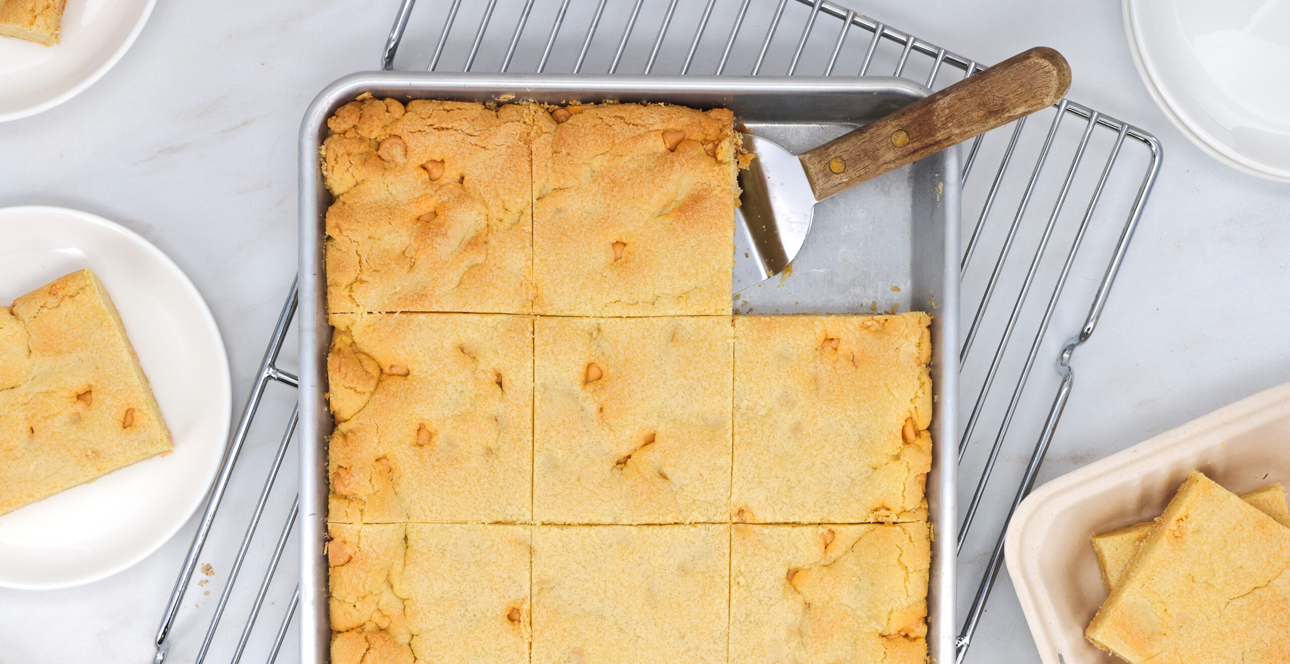 Golden brown cornbread squares arranged in a baking pan with a wooden-handled spatula nearby on a cooling rack.