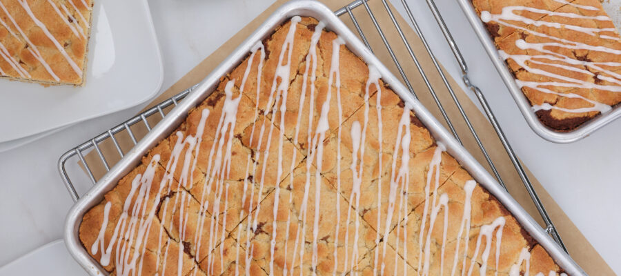 Overhead view of golden-brown lemon bars with white icing drizzled across in metal baking pans and racks.