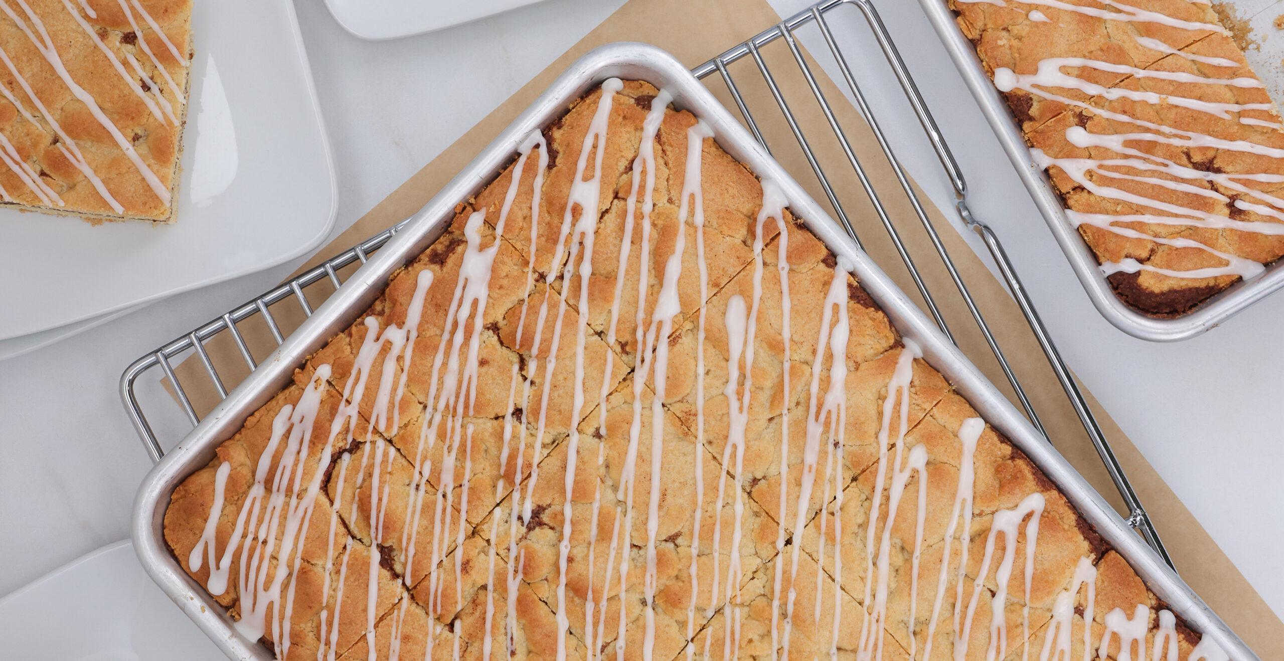 Overhead view of golden-brown lemon bars with white icing drizzled across in metal baking pans and racks.