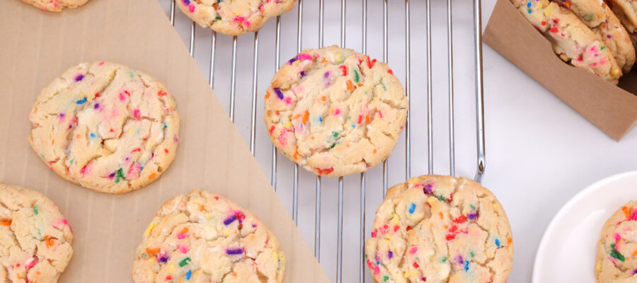 Rainbow-sprinkled sugar cookies cooling on a metal rack with parchment nearby in a kitchen setting.