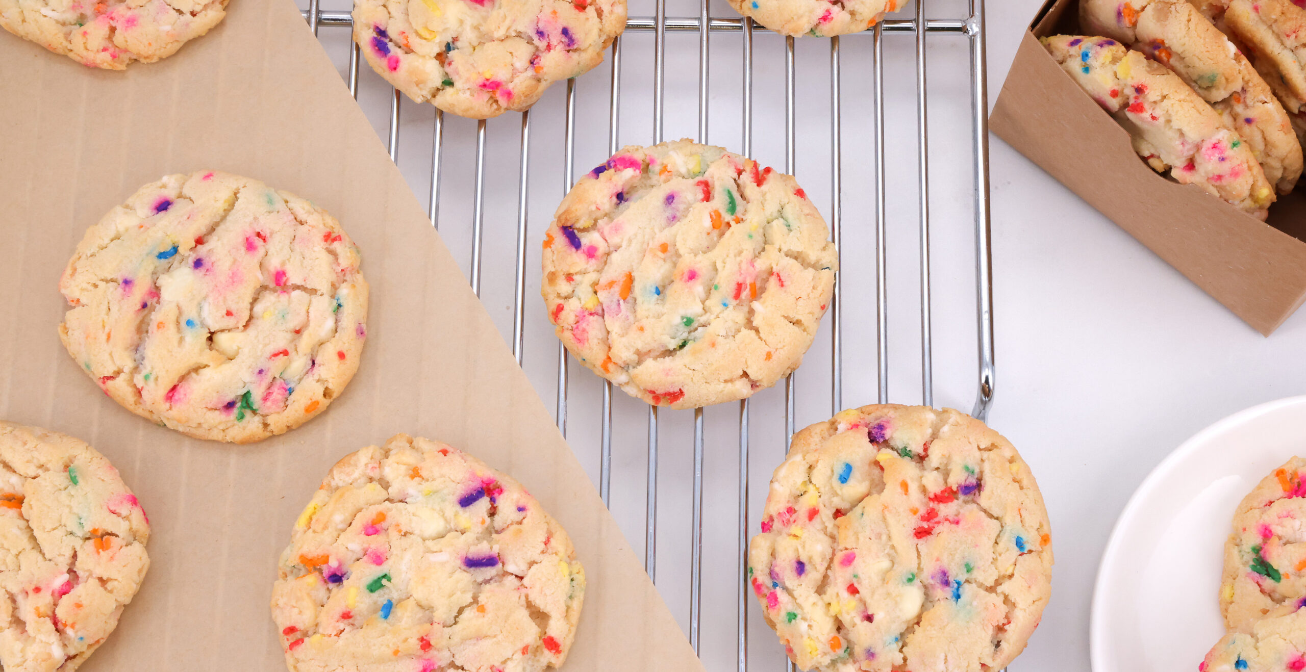 Rainbow-sprinkled sugar cookies cooling on a metal rack with parchment nearby in a kitchen setting.