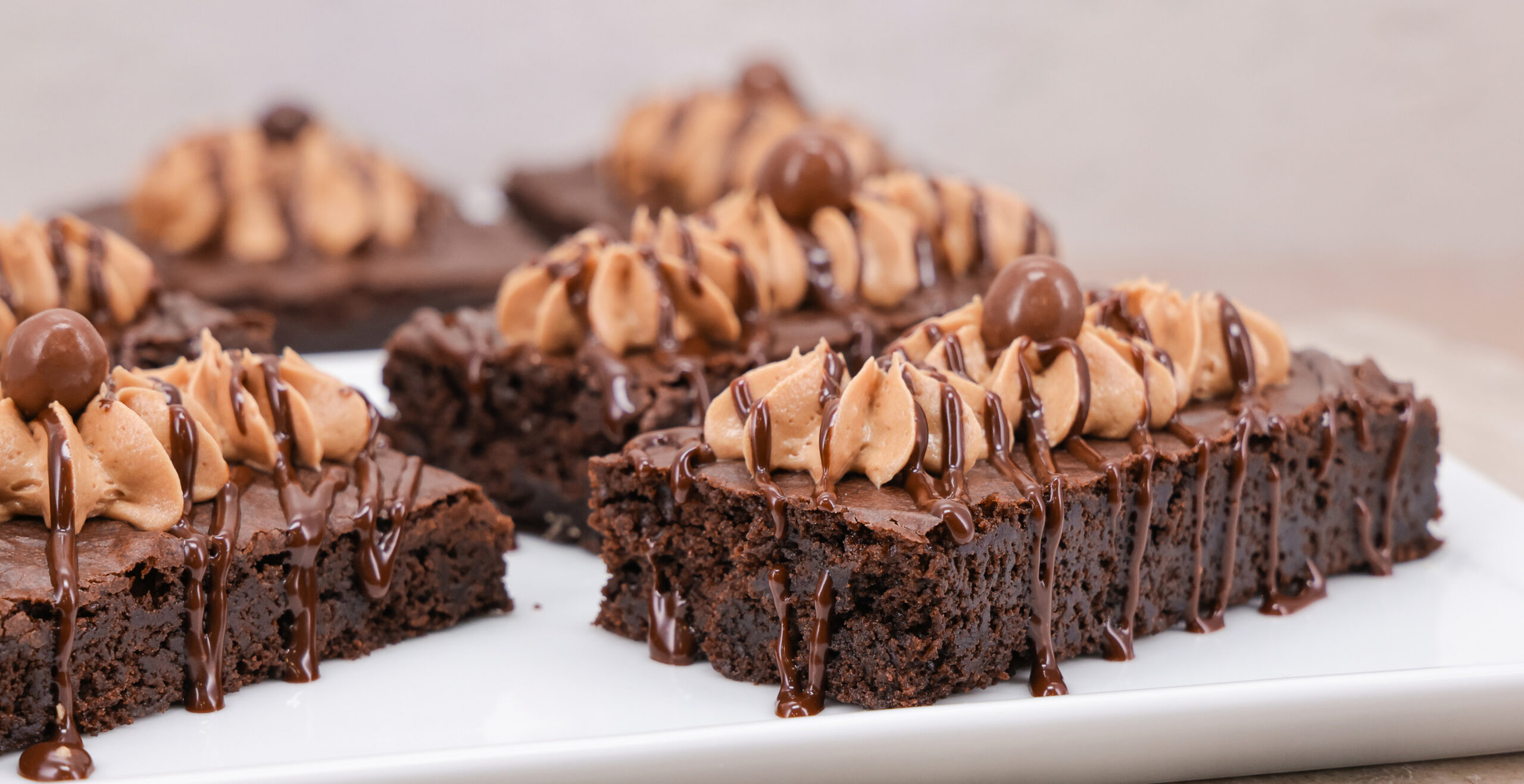 Chocolate brownies topped with swirls of peanut butter frosting and drizzled with chocolate sauce on a white plate.