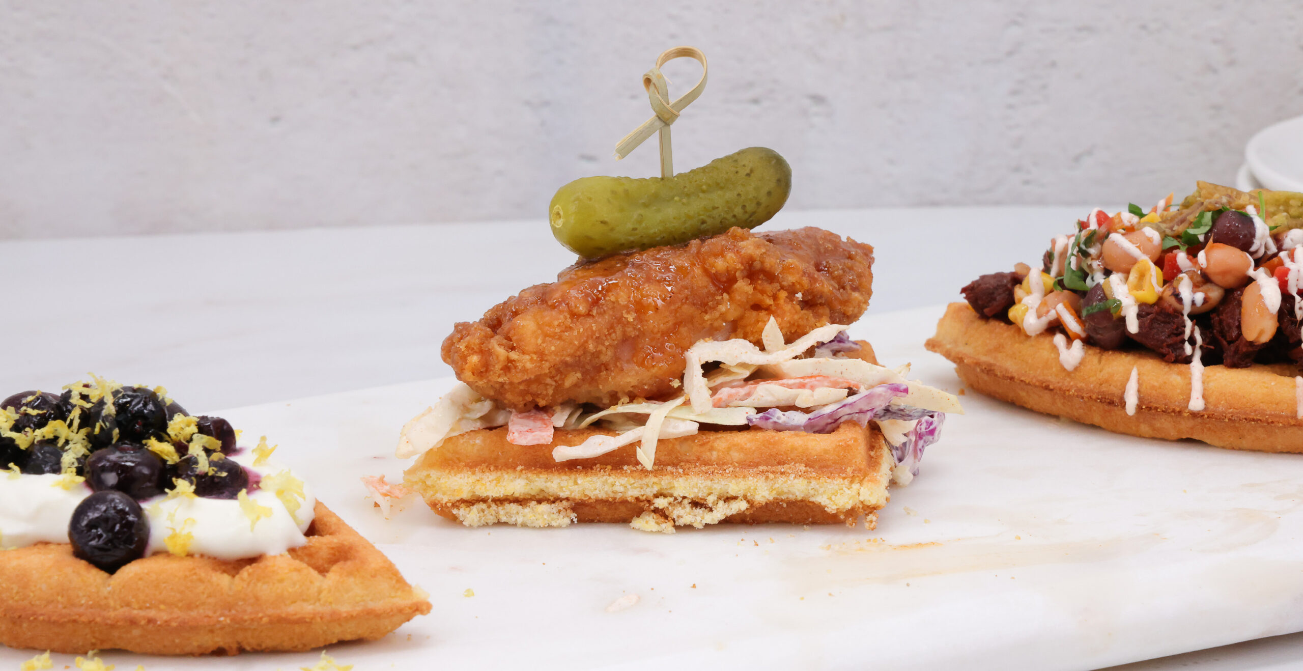 Fried chicken slider on toasted bread with coleslaw and a pickle spear, on a white surface; another loaded toast is nearby.