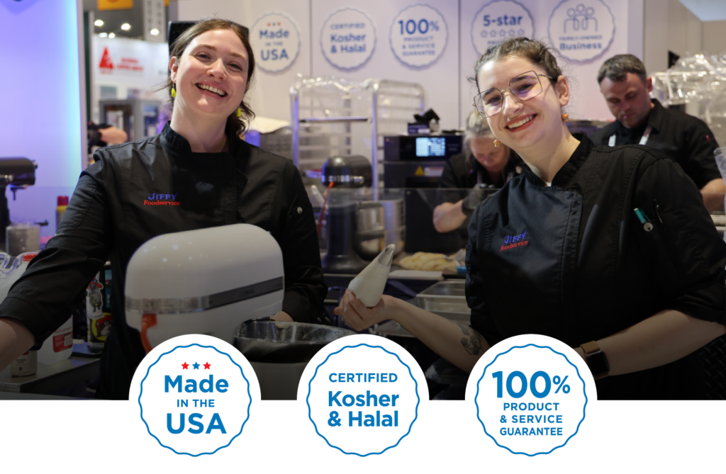 Two smiling women in black uniforms work at a busy booth, one holding a pastry bag and the other near a mixer, in a kitchen-like trade show setting.