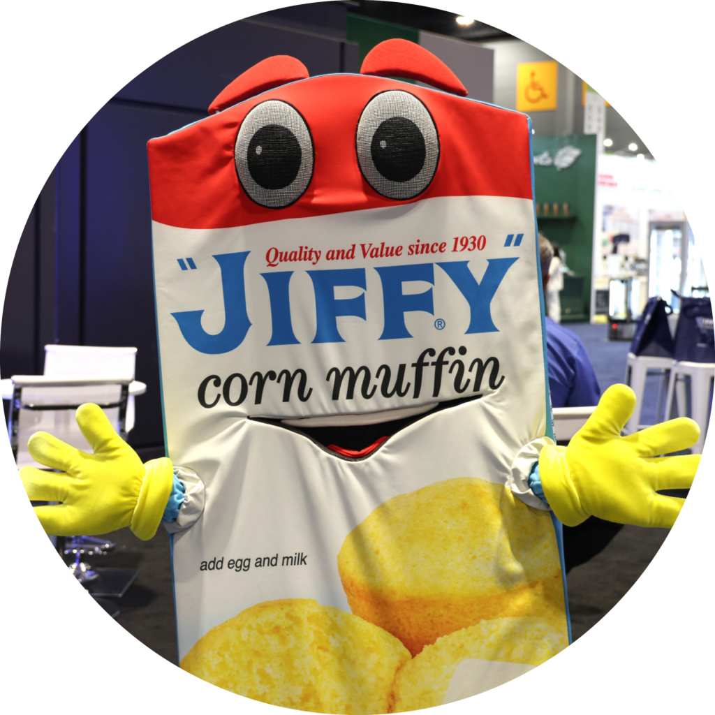 Costumed mascot with a red head and big cartoon eyes holding a banner that reads